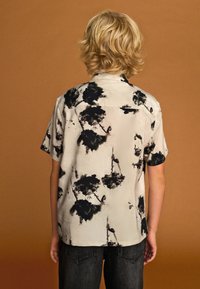 Chemise à manches courtes beige clair avec un motif floral noir, dotée d'un col à boutons et d'une texture tissée lisse.