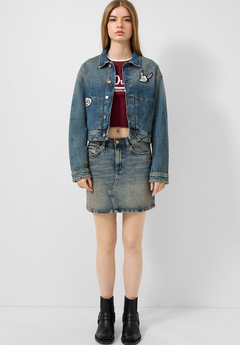 Jeansjacke mit kurzer Passform, verziert mit Applikationen, kombiniert mit einem leicht blauen Jeansrock. Schwarze Stiefeletten runden den Look ab.