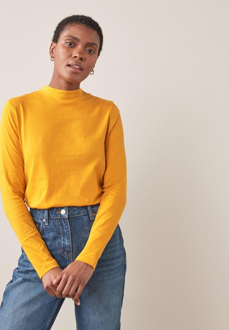 Langærmet, mock-neck-top i lys gul bomuld, parret med højtaljede, blå denim jeans med en klassisk knaplukning.