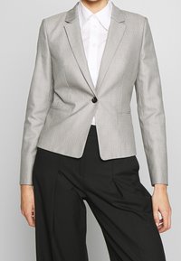Blazer gris à fines rayures avec une coupe ajustée, fermeture à un bouton, revers aiguisés et épaules structurées, porté sur une chemise blanche.