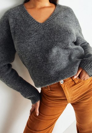 Femme portant un pull gris à col en V et un pantalon en velours côtelé orange brûlé taille haute, posant avec une main dans la poche.