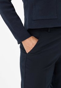 Personne portant une tenue bleu marine foncé avec une main placée de manière décontractée dans la poche avant d'un pantalon ajusté.