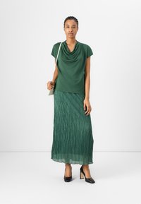 Top verde con scollo a cowl e maniche corte, abbinato a una gonna maxi verde plissettata. Scarpe col tacco alte nere e una borsa chiara.