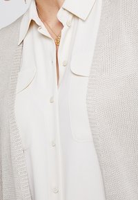 Cardigan lavorato a maglia grigio chiaro sopra una camicia bianca con bottoni. Il cardigan ha una chiusura frontale aperta ed è testurizzato, con dettagli di cucitura visibili.