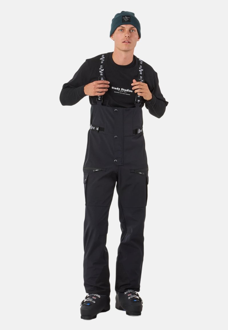 Schwarze Schneeoveralls mit verstellbaren Trägern, Druckknöpfen und Reißverschlusstaschen. Getragen über einem langen schwarzen Shirt und kombiniert mit dunklen Stiefeln.