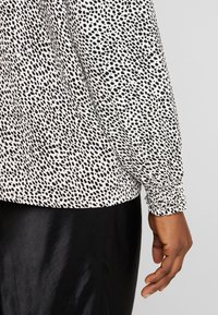 Schwarz-weiß gepunktete Bluse mit langen Ärmeln, elastischen Bündchen und einem leichten Stoff. Kombiniert mit einem schwarzen glatten Rock.