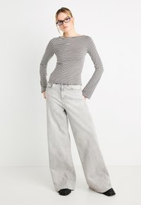 Μπλούζα με μαύρες και λευκές ρίγες και μακριά μανίκια, συνδυασμένη με ψηλόμεσα, φαρδιά τζιν παντελόνια σε ανοιχτό γκρι και μαύρα παπούτσια, σε απλό φόντο.