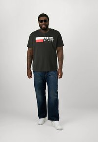 Tmavé tričko s krátkým rukávem s nápisem „JACK & JONES“, červené a bílé akcenty; spárováno s světle modrými rovnými džíny a bílými teniskami.