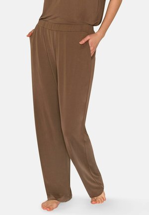 Žena stojící bosá na světlém pozadí, oblečená v volných hnědých kalhotách s elastickým pasem a bočními kapsami.