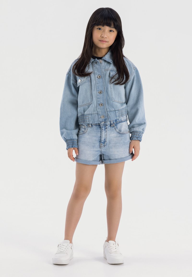 Veste en denim bleu clair avec une coupe courte, une taille élastique et un fermoir à boutons, associée à des shorts en denim bleu clair et des baskets blanches.