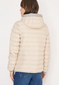 Veste matelassée beige avec motif matelassé horizontal, doublure de capuche grise et poignets côtelés. Caractérisée par une coupe décontractée et une texture douce.