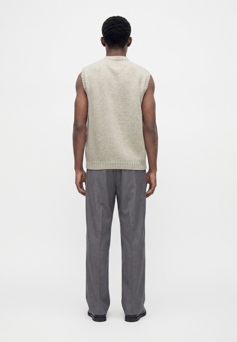 Mann von hinten gesehen, trägt einen beigen ärmellosen Pullover, graue weite Hosen und schwarze Schuhe vor einem schlichten weißen Hintergrund.