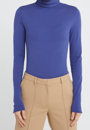 Femme portant un pull col roulé violet ajusté à manches longues et un pantalon beige taille haute avec poches latérales.