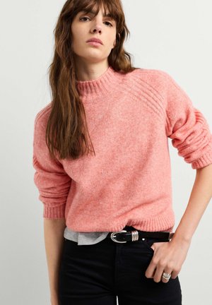 Frau mit langen braunen Haaren, die einen rosa flauschigen Pullover, schwarzen Gürtel und dunkle Hosen trägt, steht mit einer Hand in der Hüfte.