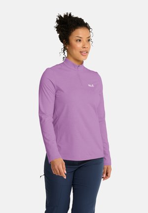 Donna con capelli ricci che indossa una maglia viola con cerniera a maniche lunghe e pantaloni blu scuro, rivolta leggermente verso destra su sfondo bianco.