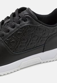 Sneaker nera con pelle texturizzata e marchi ripetuti, chiusura con lacci e suola in gomma bianca con un motivo a intaglio geometrico.