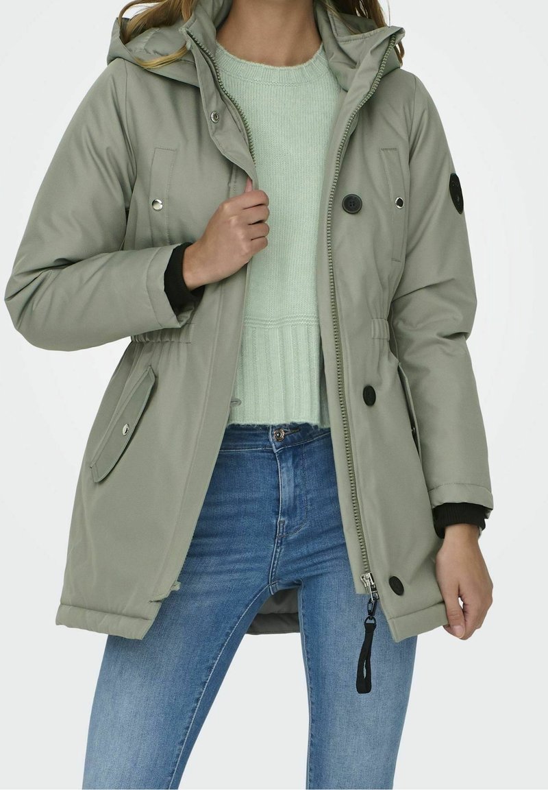Olivgrön parka med huva, framdragkedja, tryckknappar och två sidofickor. Har elastiska muddar och midjan är insvängd.