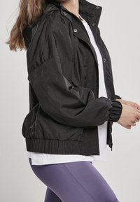 Zwarte lichtgewicht jas met een aansluitende onderrand, capuchon en elastische manchetten. Gecombineerd met een wit T-shirt en paarse leggings, van een gestructureerde stof.