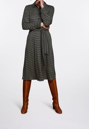 Šaty do kolen s dlouhým rukávem v tmavě zelené barvě s geometrickým vzorem; obsahují pásek na zavázání a jsou doplněny hnědými botami po kolena.