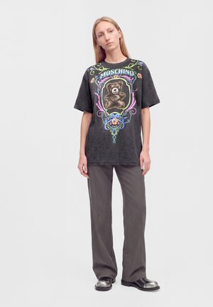 Femme aux longs cheveux blonds portant un t-shirt noir Moschino avec un motif d'ours en peluche, un pantalon gris foncé et des chaussures noires, debout sur fond blanc.