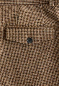Brown checkered fabric with a textured weave. Features a flap pocket and a black button. Subtle multicolored specks throughout the design.