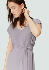 Lavendel V-Ausschnitt Kleid aus weichem Stoff, mit kurzen Ärmeln, einem Bindegürtel in der Taille und einer dezenten Textur sowie einer einzelnen Brusttasche.