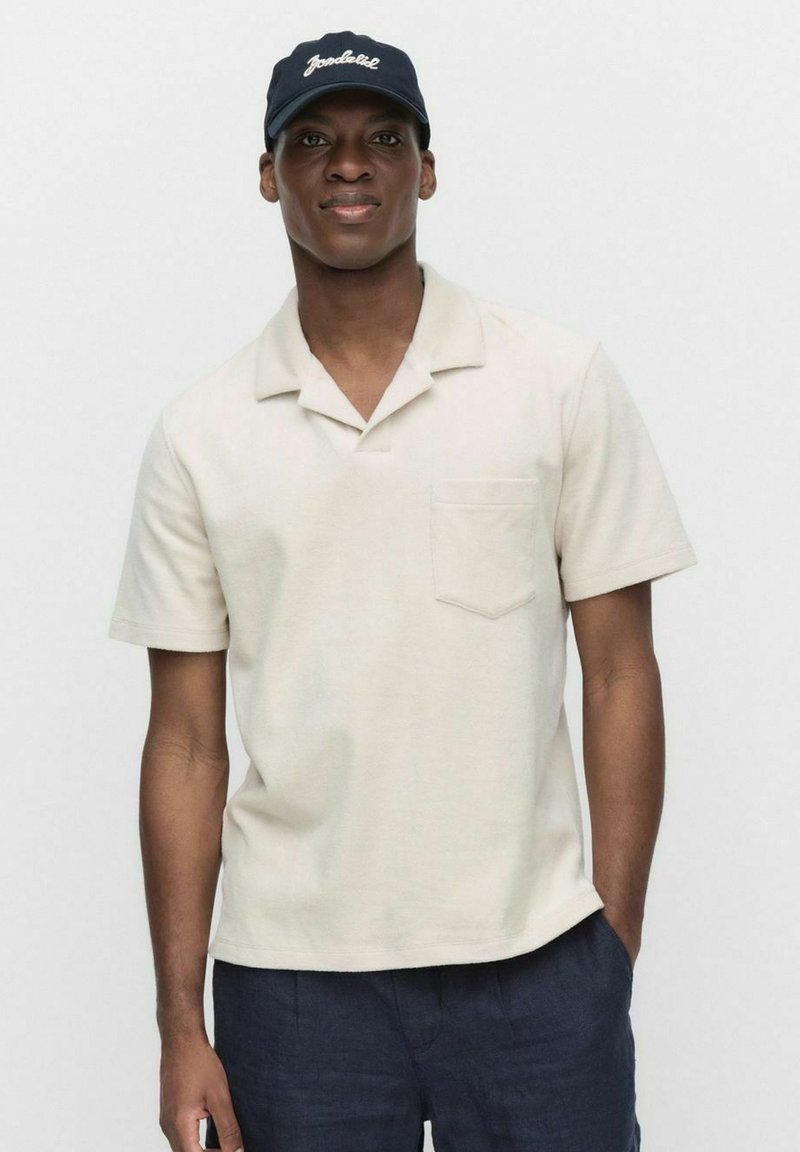 Beżowa koszulka polo z krótkim rękawem, z kieszenią na piersi i kołnierzykiem typu notch, wykonana z teksturowanej tkaniny, połączona z ciemnoniebieskimi szortami i granatową czapką.