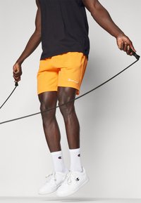 Oranžid sportlikud lühikesed püksid, millel on logo, on ühendatud valgete sokkide ja kingadega. Figuur hoiab hüppenööri, kandes musta ilma varrukateta särk.