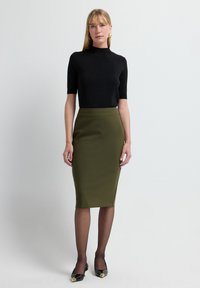 Schwarzes kurzärmliges Oberteil, hoher Ausschnitt, olivgrüner Bleistiftrock, durchsichtige schwarze Strumpfhose, schwarze Schuhe mit goldenen Akzenten, minimalistisches Design.