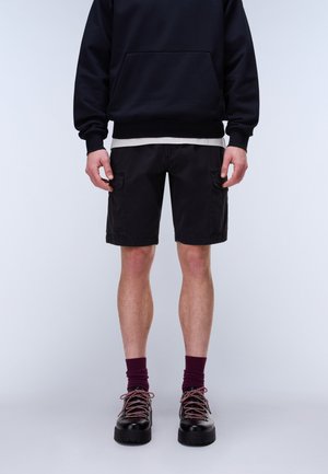 Persona con shorts cargo negros, sudadera negra, calcetines granate y botas de senderismo negras, de pie frente a un fondo claro y liso.