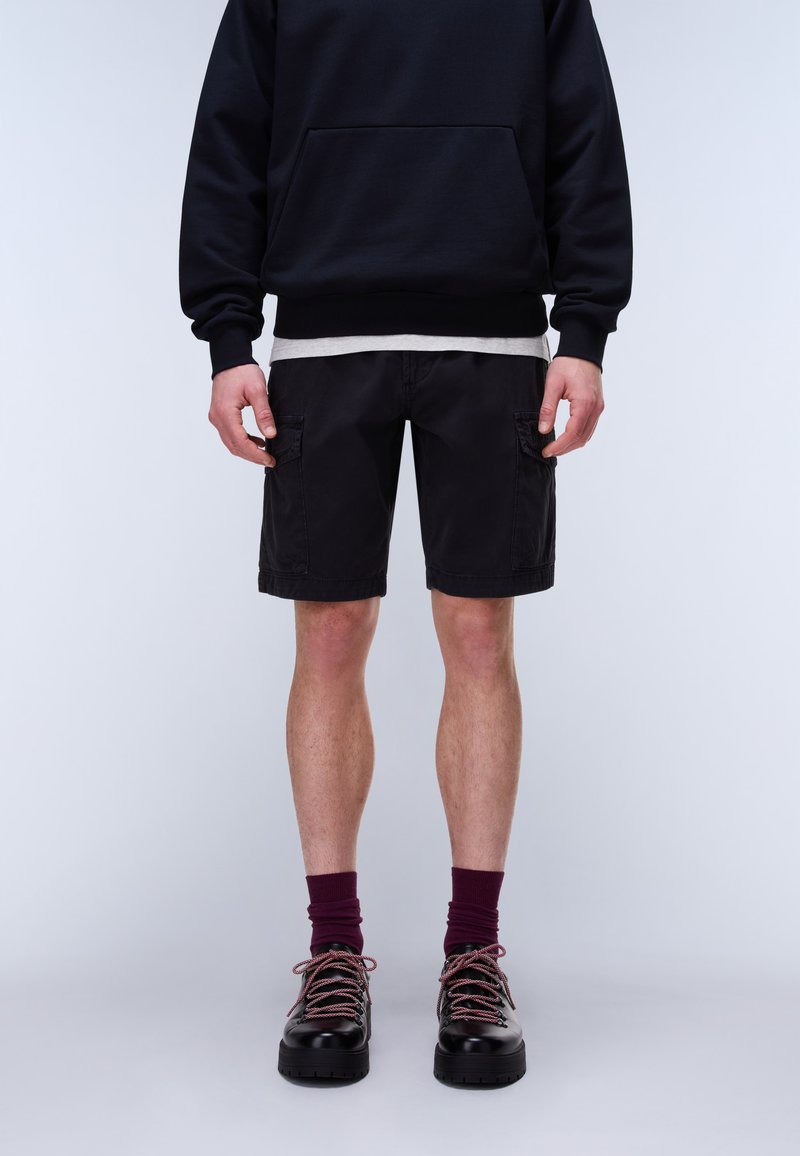 Inimene, kes kannab musti cargo lühkse, musta pusa, burgundia sokke ja musti matkasaapaid, seisab ühevärvilise heleda tausta ees.