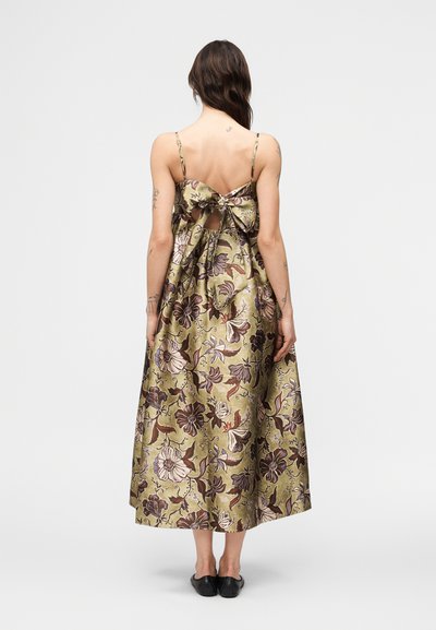 Γυναίκα που φοράει μεσοφόρεμα ελιάς με λουλούδια, με λεπτές τιράντες και ανοιχτή πλάτη δεμένη με μεγάλο φιόγκο, σε συνδυασμό με μαύρα επίπεδα παπούτσια.