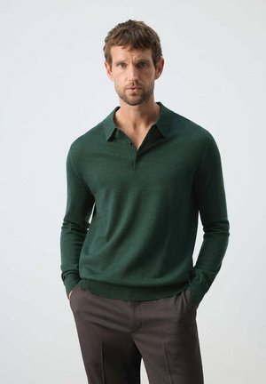 Man wearing a dark green long-sleeve polo shirt and dark trousers standing with hands in pockets against a plain background.