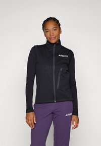 Black vest with high collar, textured pattern, and zippered pocket. Worn over a long-sleeve top, paired with purple pants. Features logo.