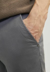 Pantalones grises texturizados con un detalle de bolsillo visible. La tela parece tejida y hay un acento de botón en el borde del bolsillo.