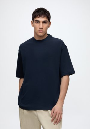 Jeune homme aux cheveux courts et foncés portant une chemise bleu marine unie à manches courtes et un pantalon beige, debout devant un fond blanc.