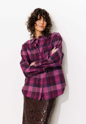 Chemise oversize à carreaux violets avec manches longues, dotée d'une poche avant et d'une fermeture à boutons, associée à une jupe à paillettes.