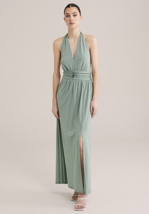 Langes, halterförmiges grünes Kleid aus schimmerndem Stoff, mit einer taillierten Taille und Knotendetail sowie einem seitlichen Schlitz; kombiniert mit Riemchensandalen.
