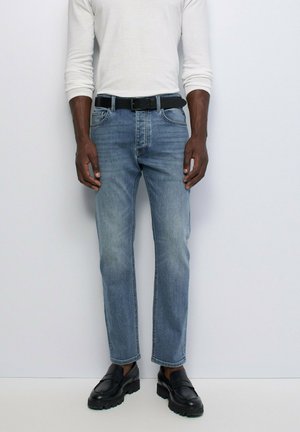 Homme portant un jean en denim bleu clair, une ceinture noire, des mocassins noirs et une chemise blanche à manches longues, debout contre un mur clair.