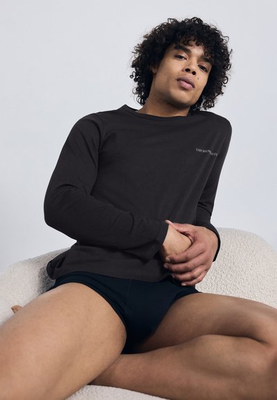 Donkerbruine, lange mouwen shirt van zachte stof, met een ronde halslijn en subtiele logodetail. Gecombineerd met donkere korte broek.