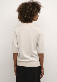 Svetlo béžový sveter s krátkymi rukávmi, vyrobený z lesklého materiálu. Má rebrovaný golier a lem, s priliehavým strihom.