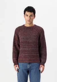 Strikkegenser i dype røde og grå nyanser, med rund hals, ribbestrikkede mansjetter og avslappet passform med et teksturert mønster. Kombinert med blå jeans.