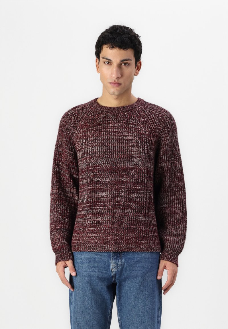 Strikkegenser i dype røde og grå nyanser, med rund hals, ribbestrikkede mansjetter og avslappet passform med et teksturert mønster. Kombinert med blå jeans.