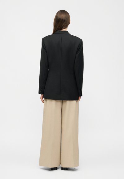 Žena s dlhými vlasmi, ktorá má na sebe čierny blejzer a široké béžové nohavice, stojaca chrbtom k divákovi pred jednoduchým bielym pozadím.