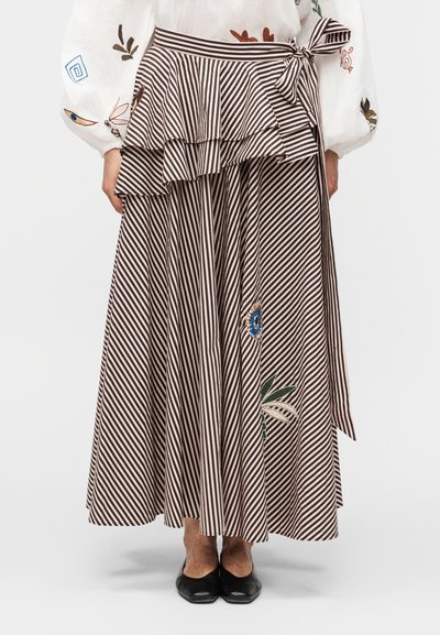 Mujer con falda en capas de rayas diagonales marrones y blancas con bordado floral, combinada con zapatos planos negros y blusa blanca bordada.