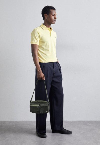 Gul polo-shirt, mørke, vide bukser og en lille grøn crossbody taske med lynlåse. Tøjet har en afslappet pasform og teksturerede materialer.