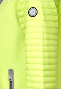 Neonově zelená bunda s texturovaným nadýchaným rukávem, obsahující kruhové logo CECIL a zipovou kapsu v kontrastní šedé barvě.