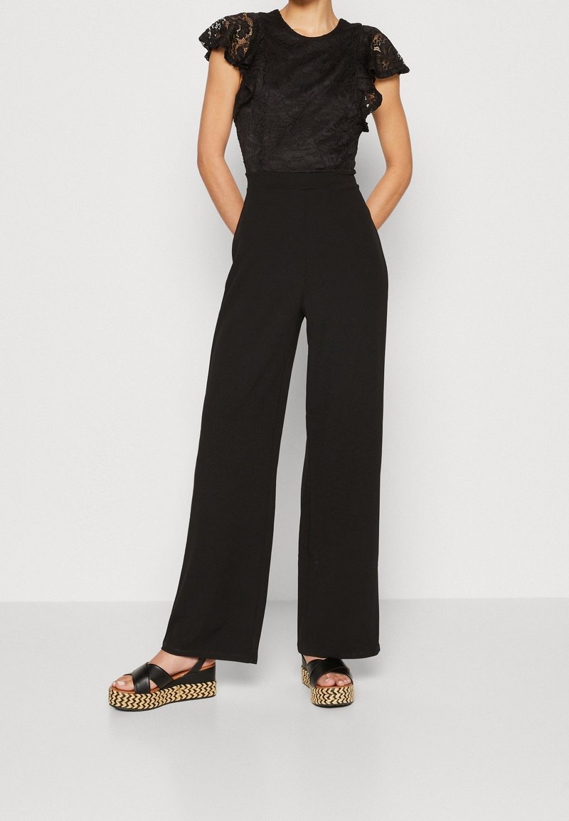 Personne portant un haut court à manches courtes en dentelle noire, un pantalon large noir taille haute et des sandales compensées noires avec des semelles tissées.