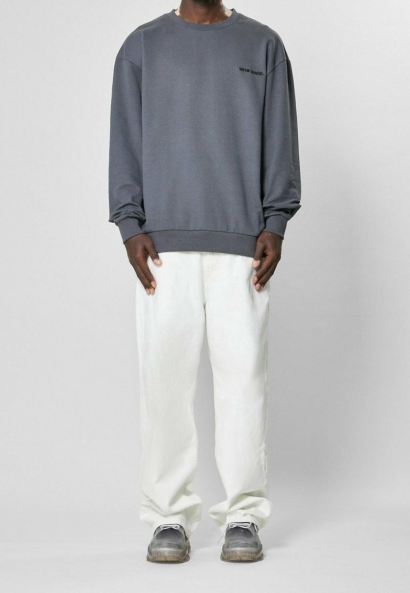 Felpa grigia con scollo rotondo e polsini a costine, abbinata a pantaloni chiari e comodi. Sneakers con suola trasparente completano l'outfit.