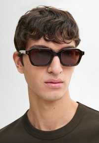 Occhiali da sole tortoiseshell neri e marroni con forma quadrata, lenti scure e dettagli laterali trasparenti, indossati da una persona con capelli corti.